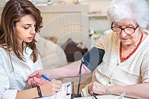 Nurse measuring blood pressure of senior woman at home. Female doctor writing medical results to clipboard.