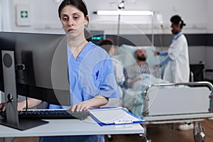 Nurse at medical desk typing patient data into computer while team of doctors putting iv drip line