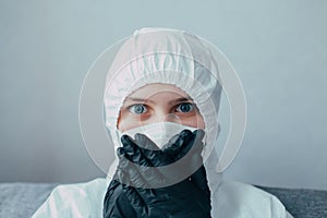 Nurse in medical mask and viral protective suit covers her face with black gloves on hands in quarantine due to the consequences o