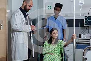 Nurse moving to treatment room woman with illness sitting in wheelchair holding drip pole