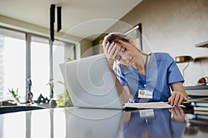 Nurse paying bills online, sitting in living room, working on notebook. Doctor in scrubs dealing with work issues from