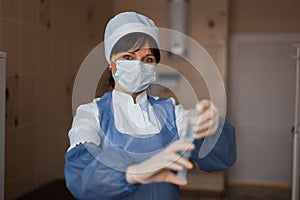 Nurse in a protective mask in medical clothes is standing and looking at the camera in the arm, holding a syringe in her hand.