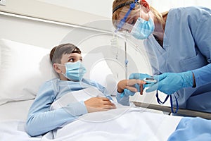 Nurse with pulse oximeter on patient child in hospital bed, wearing protective visor mask, corona virus covid 19 protection