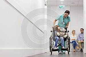 Nurse pushing boy with teddybear on wheelchair while patients waiting in hospital corridor