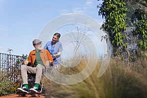 Nurse pushing senior man in wheelchair. Female caregiver and elderly man enjoying a warm autumn day in nursing home.