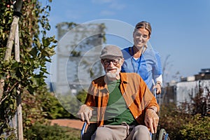 Nurse pushing senior man in wheelchair. Female caregiver and elderly man enjoying a warm autumn day in nursing home.
