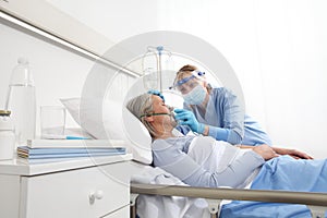 Nurse puts oxygen mask on elderly woman patient lying in the hospital room bed, wearing protective gloves and visor medical mask, photo