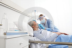 Nurse puts oxygen mask on elderly woman patient lying in the hospital room bed, wearing protective gloves and visor medical mask,