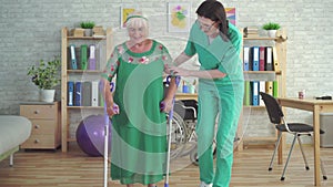 Nurse in rehabilitation clinic supports elderly woman on crutches