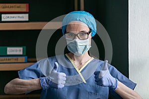 Nurse shows thumbs up sign giving positive prognosis. Female medic in blue protective uniform and disposable gloves