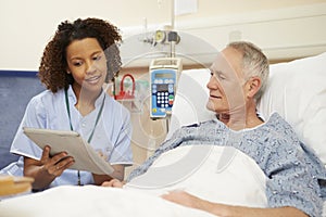 Nurse Sitting By Male Patient's Bed Using Digital Tablet