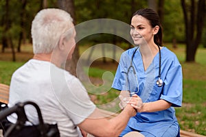 A nurse is smiling and holding the hand of an old man who is sitting next to in a wheelchair in the park