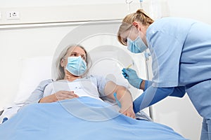 Nurse with the syringe injects the vaccine to the elderly woman patient lying in the hospital room bed, wearing protective gloves