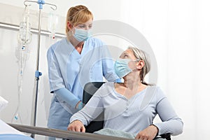 Nurse take comfort elderly woman in wheelchair wearing surgical protective medical masks in hospital room, concept of isolation