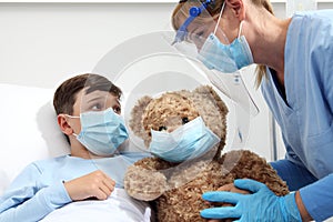 Nurse takes care of the patient child in hospital bed playing with teddy bear, wearing protective masks, corona virus covid 19