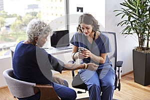 Nurse Wearing Scrubs In Office Checking Senior Female Patients Blood Pressure