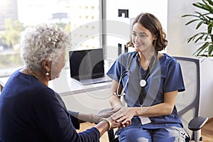 Nurse Wearing Scrubs In Office Reassuring Senior Female Patient And Holding Her Hands