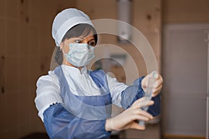 Nurse in a white mask in blue medical clothing stands holding a syringe in her hand.