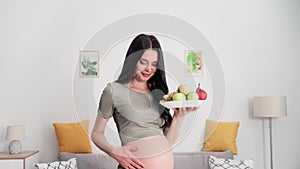 nutrition pregnancy, beautiful young woman strokes big belly with plate of healthy tasty fruits while standing in cozy