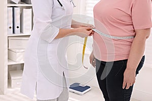 Nutritionist measuring overweight woman`s waist with tape in clinic, closeup