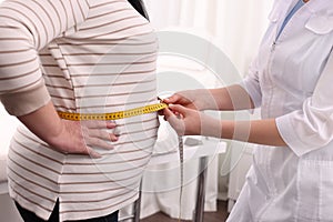 Nutritionist measuring overweight woman`s waist with tape in clinic, closeup