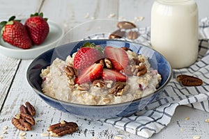 Nutritious oatmeal bowl with fresh strawberries, nuts and milk on white wooden table