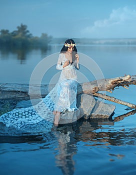 A nymph with long dark hair in a white vintage dress sits on a log across the river. in the hair a wreath of lilies
