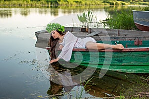 The nymph with long dark hair in a white vintage dress sitting in a boat in the middle of the river.