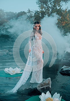 The nymph with long dark hair in a white vintage dress stands on a stone in the middle of the river. in the hair a
