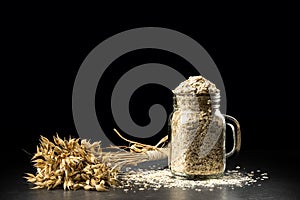 Oat bunch and flakes in flavouring jar, on black background. Grain bouquet, golden oats spikelets on dark wooden table, c