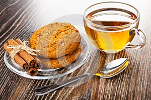 Oat cookies with sunflower seeds, cinnamon sticks in saucer, cup with tea, spoon on wooden table