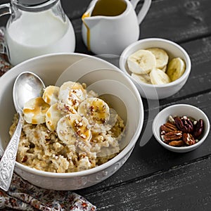 Oatmeal with banana, caramel sauce and pecan nuts in a white bowl on a dark wooden surface