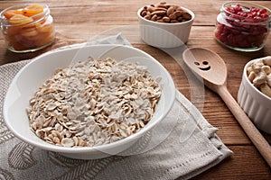 Oat flakes in bowl with dried apricots, dried cherries, almonds, cashews in jars on wooden table with wooden spoon.