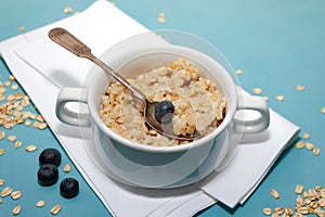 Oatmeal porridge with fresh blueberries in white bowl on blue background, linen napkin, silver spoon