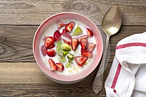 Oatmeal porridge with slices of kiwi, strawberries in pink bowl, spoon, napkin with red stripes on wooden background Top view Flat
