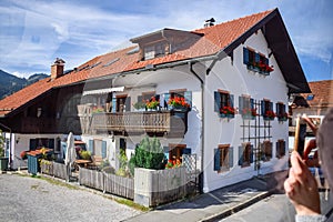 OBERAMMERGAU, GERMANY - OKTOBER 09, 2018: A tourist photographs on phone house with red flowers on balcony