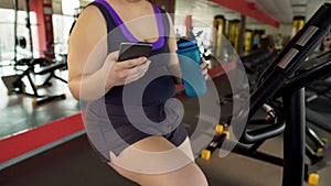 Obese female sitting on exercise bike, drinking water and scrolling on cellphone