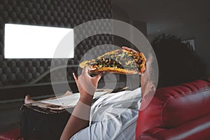 Obese man eats pizza in front of a TV at night