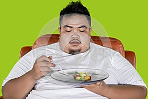 Obese man looks hesitate to eat salad on studio photo