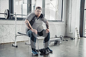 Obese man sitting on bench and Looking At Camera after exercising with dumbbells at gym