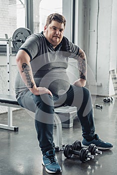 Obese man sitting on bench and Looking At Camera after exercising with dumbbells at gym