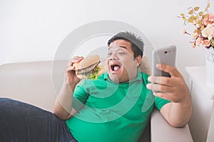 Obese person eating hamburger while using mobile phone