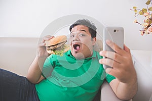 Obese person eating hamburger while using mobile phone