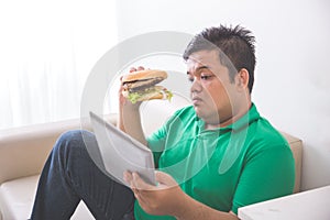 Obese person eating hamburger while using tablet pc