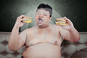 Obese person eats cheeseburger on sofa