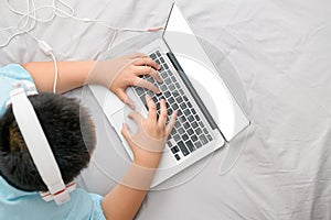 Obese schoolboy lying and wearing headphones to learn online from a laptop on bed