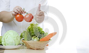 Obese woman holding fresh tomatoes and showing thumps up with non-toxic vegetables on marble table in white kitchen