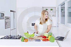 Obese woman making salad while looking at laptop