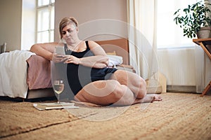 Obese woman in underwear relaxing at home, sitting, using smartphone and drinking wine.