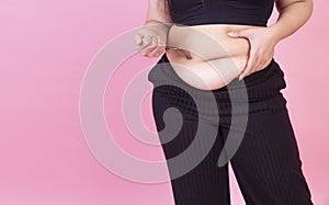 Obesity unhealthy weight loss concept. Hand of fat woman holding fork on her belly while standing over isolated pink background.